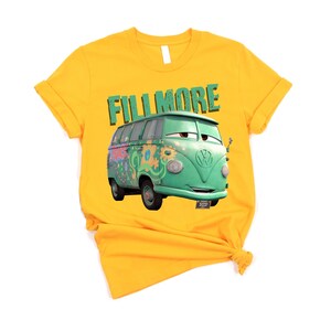May include: Mustard yellow t-shirt featuring a cartoon image of Fillmore, a green VW bus from the movie Cars. The word "FILLMORE" is printed above the bus in green block letters. The shirt is knotted at the bottom.