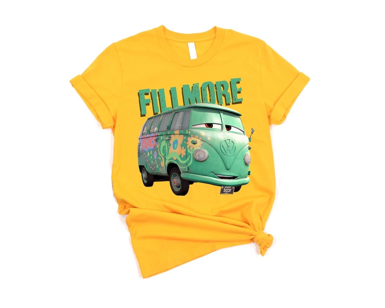 May include: Mustard yellow t-shirt featuring a cartoon image of Fillmore, a green VW bus from the movie Cars. The word "FILLMORE" is printed above the bus in green block letters. The shirt is knotted at the bottom.