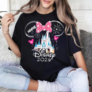 Op de afbeelding: Zwart T-shirt met een Disney kasteelafbeelding, roze strik, vuurwerk en ballonnen. Het shirt heeft de tekst "Disney 2026" en ruimte voor een naam. Het ontwerp is in cartoonstijl.