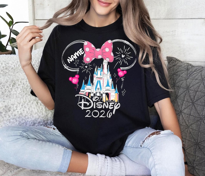 Op de afbeelding: Zwart T-shirt met een Disney kasteelafbeelding, roze strik, vuurwerk en ballonnen. Het shirt heeft de tekst "Disney 2026" en ruimte voor een naam. Het ontwerp is in cartoonstijl.