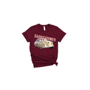 May include: A maroon t-shirt featuring a cartoon car with the text "BARNSTORMER" above it. The car is beige with a pink visor and the number "84" on the side. The shirt has a crew neck and short sleeves.
