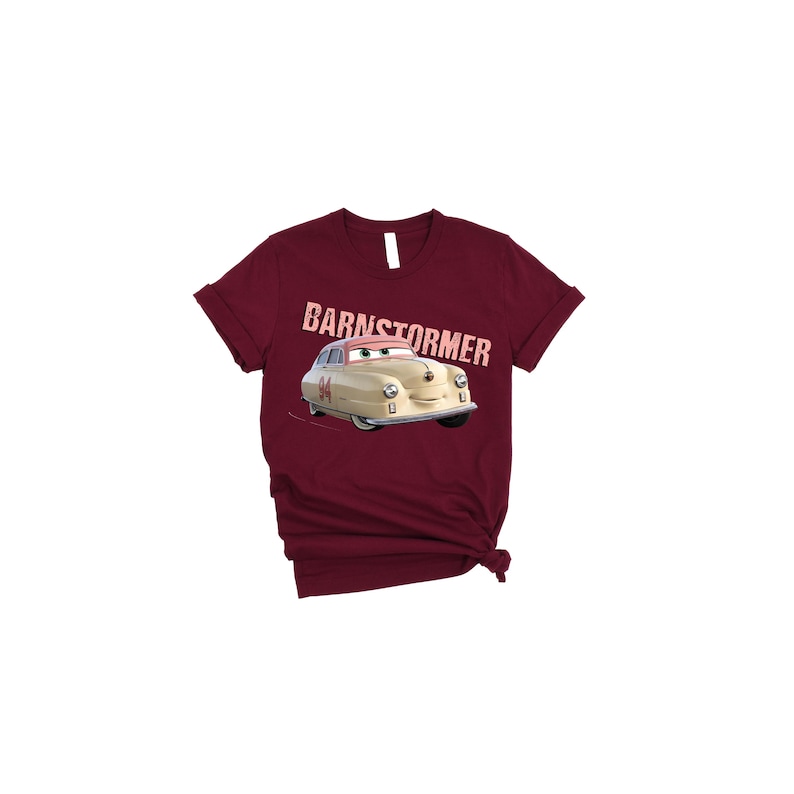 May include: A maroon t-shirt featuring a cartoon car with the text "BARNSTORMER" above it. The car is beige with a pink visor and the number "84" on the side. The shirt has a crew neck and short sleeves.