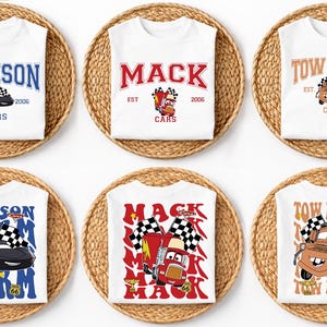 May include: White t-shirts with various Cars movie designs. Designs include the names "Jackson", "Mack", and "Tow Mater", along with car illustrations and the text "Cars" and "EST 2006". The shirts are displayed on woven mats.