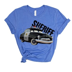 May include: A light blue t-shirt featuring a cartoon image of a black and white sheriff car with the word "SHERIFF" in black. The car has a red light on top and the word "SHERIFF" is printed in a diagonal font.