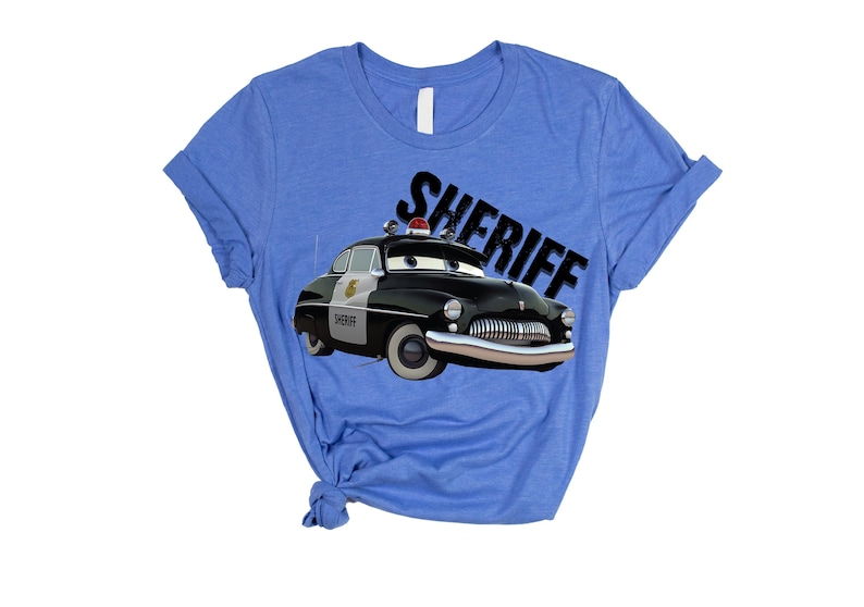 May include: A light blue t-shirt featuring a cartoon image of a black and white sheriff car with the word "SHERIFF" in black. The car has a red light on top and the word "SHERIFF" is printed in a diagonal font.