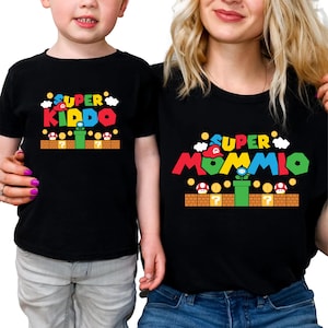 May include: Black t-shirts with matching graphics. One shirt says "Super Kiddo" and the other says "Super Mommio" in colorful text. The design includes a green pipe, question mark blocks, and mushroom details, resembling a video game theme.