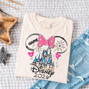 Op de afbeelding: Cr&egrave;mekleurig T-shirt met een Disney kasteelontwerp, een roze strik met stippen, Mickey Mouse oren en de tekst "Disney 2026". Het shirt wordt getoond met een blauwe jeans.