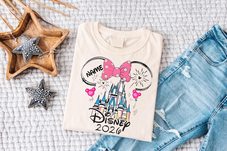 Op de afbeelding: Cr&egrave;mekleurig T-shirt met een Disney kasteelontwerp, een roze strik met stippen, Mickey Mouse oren en de tekst "Disney 2026". Het shirt wordt getoond met een blauwe jeans.