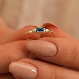 May include: A delicate gold ring featuring a rectangular, dark blue gemstone as the centerpiece, flanked by two smaller, round, clear gemstones. The ring is held up against a soft, neutral background.