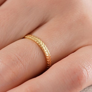 Könnte beinhalten: Ein goldfarbener Ring mit einem geflochtenen oder geflochtenen Design. Der Ring wird an einem Finger getragen und zeigt seine komplizierte Textur und den warmen metallischen Farbton. Das Design des Rings ähnelt einer Reihe von verbundenen, strukturierten Segmenten.