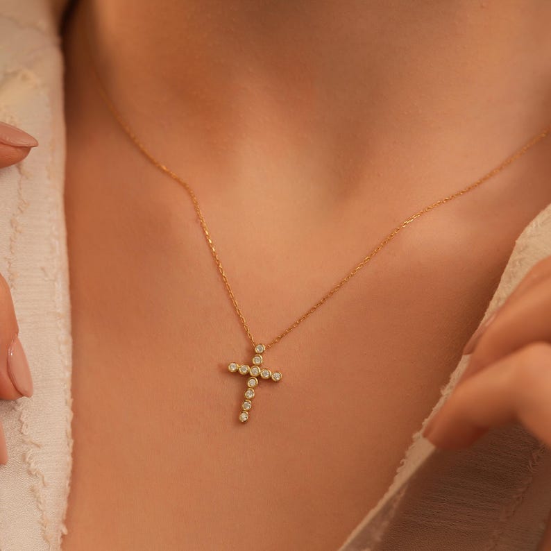K&ouml;nnte beinhalten: Eine goldfarbene Halskette mit einem Kreuzanh&auml;nger, der mit kleinen, runden, klaren Edelsteinen besetzt ist. Das Kreuz h&auml;ngt an einer feinen Kette. Die Halskette wird vor einem neutralen Hintergrund gezeigt.