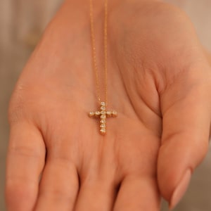K&ouml;nnte beinhalten: Eine goldfarbene Halskette mit einem Kreuzanh&auml;nger. Das Kreuz ist mit kleinen, runden, klaren Edelsteinen besetzt. Die Halskette wird in einer offenen Hand gehalten, wodurch das filigrane Design und die Handwerkskunst des Schmucks hervorgehoben werden.