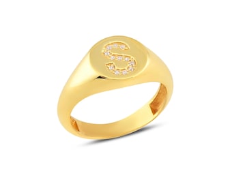 May include: A gold signet ring with a rounded face. The face of the ring features the letter "S" encrusted with small, clear gemstones. The ring has a polished, shiny finish and is set against a white background.