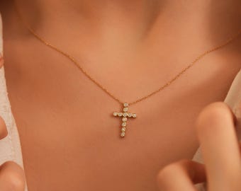 Könnte beinhalten: Eine goldfarbene Halskette mit einem Kreuzanhänger. Das Kreuz ist mit kleinen, runden, klaren Edelsteinen besetzt. Die Halskette wird um den Hals getragen, wobei das Kreuz auf der Brust liegt. Der Hintergrund ist in einer neutralen Farbe gehalten.