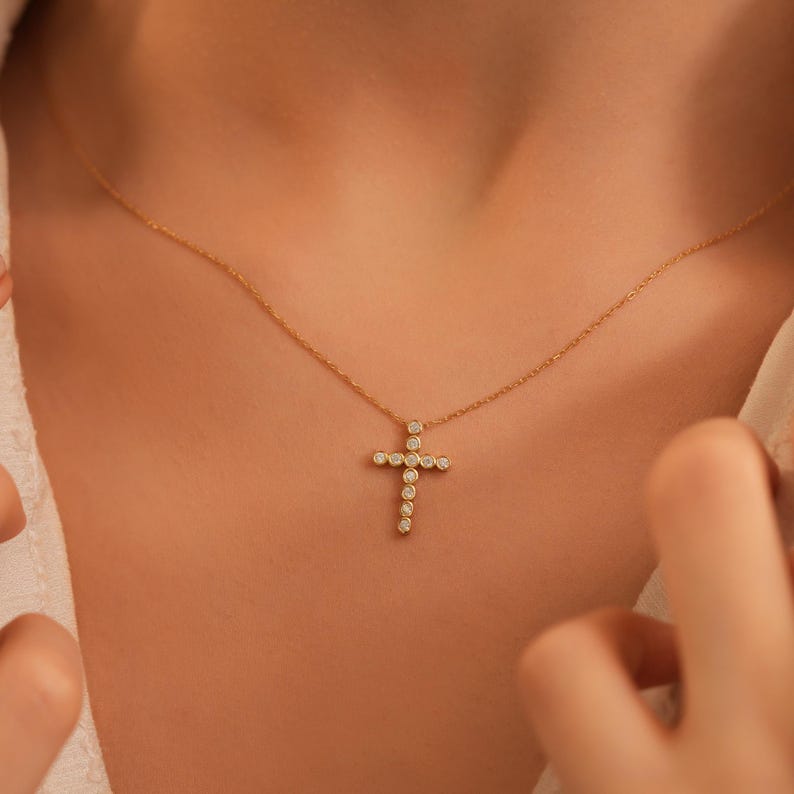 K&ouml;nnte beinhalten: Eine goldfarbene Halskette mit einem Kreuzanh&auml;nger. Das Kreuz ist mit kleinen, runden, klaren Edelsteinen besetzt. Die Halskette wird um den Hals getragen, wobei das Kreuz auf der Brust liegt. Der Hintergrund ist in einer neutralen Farbe gehalten.