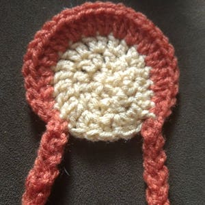 May include: A handmade crochet item in a circular shape with two braided strands. The center is cream-colored, surrounded by a rust-colored border. The braids and border are the same rust color. The item is likely a decorative applique or embellishment.