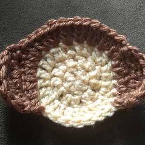 May include: A handmade crochet coaster in a half-circle shape. The coaster features a color block design with a light beige center and brown edges. The yarn is tightly woven, creating a textured surface. The coaster is designed to protect surfaces from hot or cold beverages.