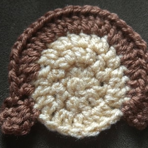 May include: A crocheted brown and beige hair piece with a rounded shape. The hair piece is made of yarn and has a textured surface.