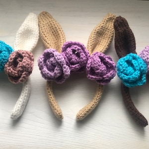 May include: Four crocheted bunny ear headbands with different colored flowers. The headbands are white, beige, brown, and feature gray, purple, blue, and pink flowers.