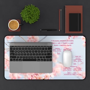 Puede incluir: Una configuración de escritorio con una computadora portátil, un ratón y una alfombrilla de ratón con estampado floral con términos contables. Una taza de café, una planta pequeña, un cuaderno y lápices también están sobre la superficie oscura. La alfombrilla del ratón tiene el texto "T-Accounts, and Debits & Credits".