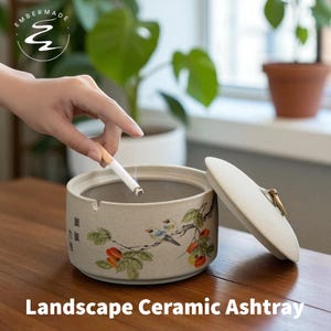 May include: A beige ceramic ashtray with a lid, featuring a painted landscape design of birds and branches. The text "Landscape Ceramic Ashtray" is at the bottom. A lit cigarette is being placed into the ashtray.