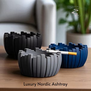 Può includere: Tre posacenere moderni neri, grigi e blu, con un design geometrico. Il posacenere grigio contiene una sigaretta accesa. Il testo "Luxury Nordic Ashtray" è in basso nell'immagine.