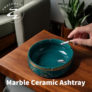 May include: A teal marble ceramic ashtray with a gold Greek key design around the rim. The ashtray is round and sits on a wooden table. The text "Marble Ceramic Ashtray" is at the bottom of the image.