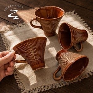 May include: Four brown ceramic mugs with a flared rim and a textured, vertical ribbed pattern on the exterior. Each mug features a curved handle and a glossy finish. The text EMBERMADE is visible in the upper left corner of the image.