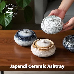 May include: A collection of Japandi ceramic ashtrays in various colors, including white speckled, navy blue, and tan. Each ashtray has a matching lid with a small knob. The ashtrays are displayed on a dark wooden table.
