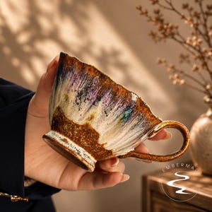 May include: A ceramic teacup with a textured, uneven rim and a handle. The exterior features a reactive glaze with streaks of deep brown, cream, purple, and blue. The base is a dark, earthy brown. The text EMBERMADE is printed with a logo.