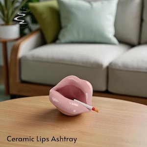 Puede incluir: Un cenicero de cerámica rosa con forma de labios, con un cigarrillo en la abertura. El cenicero está sobre una mesa de madera. El texto "Ceramic Lips Ashtray" está en la parte inferior de la imagen.
