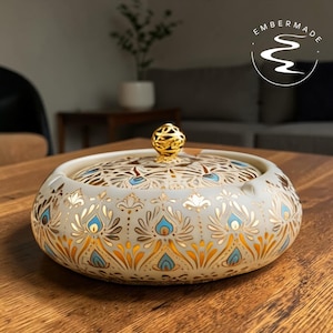 May include: A decorative ceramic ashtray with a lid. The ashtray is off-white with a gold, blue, and orange floral pattern. The lid has a gold knob. The brand logo "Embermade" is in the top right corner.