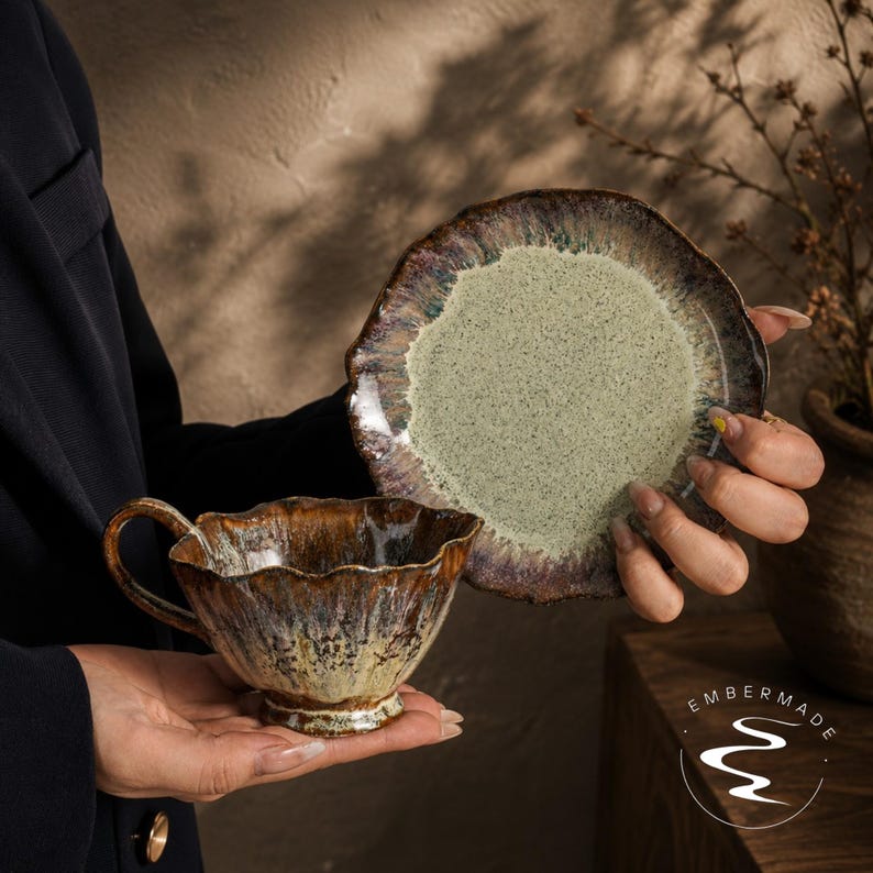 May include: A ceramic teacup and matching saucer set featuring a reactive glaze with a speckled light green center and a dark, wavy, brown-toned rim. The teacup has a scalloped edge and a curved handle, while the saucer has a matching wavy border.