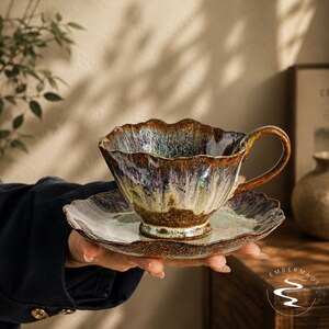 May include: A ceramic teacup and saucer set featuring a wavy, scalloped rim. The glaze displays a reactive blend of deep brown, purple, and cream tones with a glossy finish. The cup has a curved handle and sits on a matching, wide-rimmed saucer.