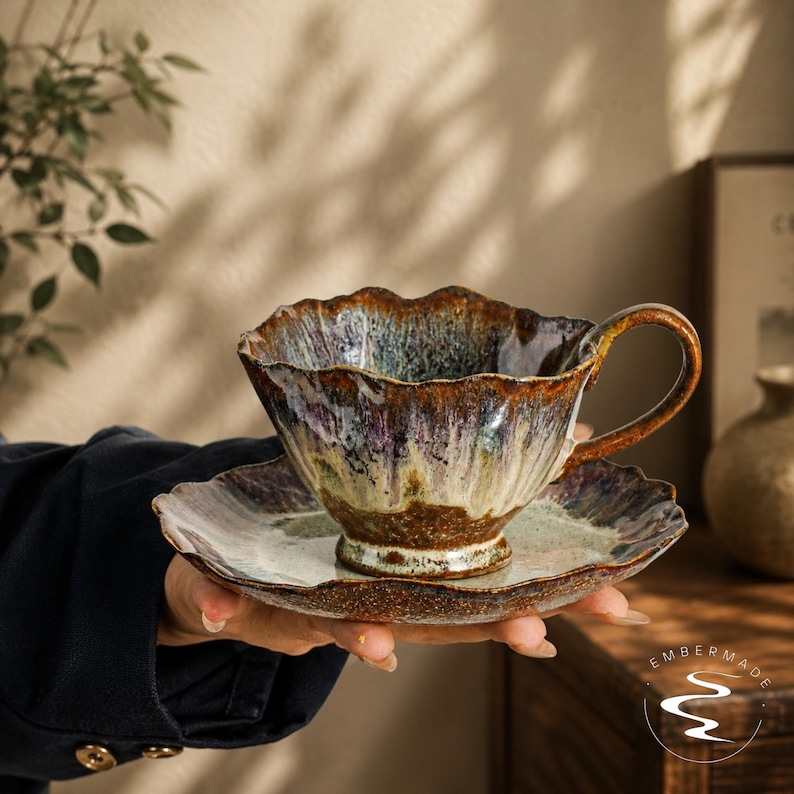 May include: A ceramic teacup and saucer set featuring a wavy, scalloped rim. The glaze displays a reactive blend of deep brown, purple, and cream tones with a glossy finish. The cup has a curved handle and sits on a matching, wide-rimmed saucer.