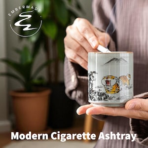 May include: A cylindrical ashtray with a gray background and a gold rim. The ashtray features a painted design of two tigers and a mountain landscape. The text "Modern Cigarette Ashtray" is at the bottom.