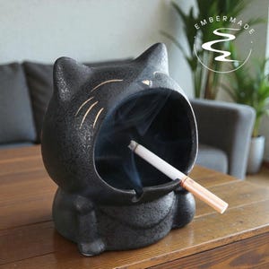 以下が含まれることがあります： 猫の頭の形をした黒いセラミック灰皿。灰皿の中にはタバコが置かれています。灰皿はテクスチャ加工された表面と金色のアクセントが特徴です。背景にはブランド名「EMBERMADE」が見えます。