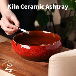 May include: A round, red ceramic ashtray with a glossy finish. The ashtray has a wide opening and cigarette rests. The text "Kiln Ceramic Ashtray" is displayed at the top of the image. The brand name "Embermade" is displayed at the bottom left.