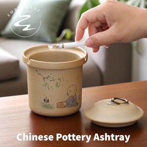 May include: Tan ceramic ashtray with a lid, featuring a hand-painted illustration of a monk. The ashtray has a cylindrical shape with a small groove for resting a cigarette. The lid is round with a metal handle. The text "Chinese Pottery Ashtray" is at the bottom.