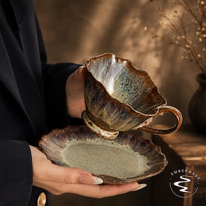Peut inclure: Un ensemble tasse et soucoupe en céramique avec un bord festonné et ondulé. L'émail présente une finition réactive et tachetée dans des tons de brun profond, bronze et bleu-vert sourd. La soucoupe a un centre moucheté et la tasse a une anse.