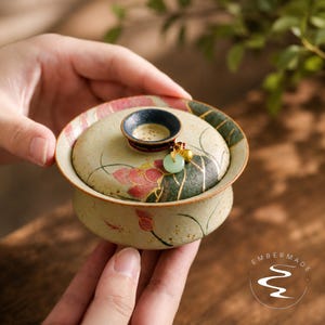 May include: A ceramic tea bowl with a lid, featuring a beige base with painted pink lotus flowers and green leaves. The lid has a dark blue knob adorned with a small gold-toned charm and a light green jade-like ring. The text Embermade is visible.
