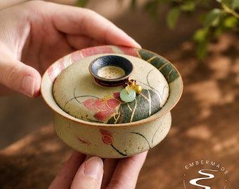 May include: A ceramic tea bowl with a lid, featuring a beige base with painted pink lotus flowers and green leaves. The lid has a dark blue knob adorned with a small gold-toned charm and a light green jade-like ring. The text Embermade is visible.