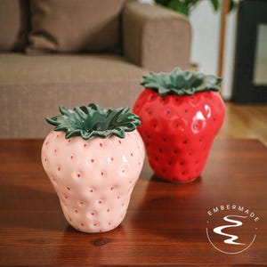 May include: Two ceramic strawberry-shaped vases. One is light pink with red speckles, the other is bright red. Both have green leafy tops. The vases are on a dark wooden surface, with a couch in the background.