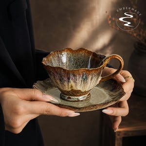 May include: A ceramic teacup and saucer set featuring a scalloped edge and a reactive glaze in shades of brown, cream, and hints of purple. The cup has a curved handle and a pedestal base. The text "EMBERMADE" is visible in the upper right corner.