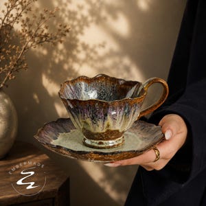 May include: A ceramic teacup and saucer set featuring a wavy, organic rim and a reactive glaze in shades of deep brown, purple, and cream. The surface has a textured, mottled appearance with a glossy finish. The logo Embermade is visible.
