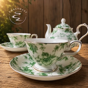 Peut inclure: Un service à thé en porcelaine blanche orné d'un motif floral vert et d'un liseré doré. L'ensemble comprend une théière, deux tasses et deux soucoupes. Le logo Embermade avec un graphique de fumée stylisé est visible en haut à gauche.