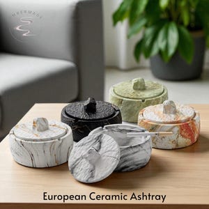 Peut inclure: Plusieurs cendriers européens en céramique de différentes couleurs et motifs. Les cendriers sont ronds avec des couvercles et un aspect texturé semblable au marbre. Un cendrier contient une cigarette. Le texte "European Ceramic Ashtray" est en bas.