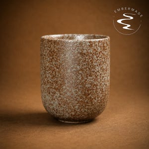 May include: A cylindrical ceramic cup with a speckled brown and off-white textured finish. The surface features a mottled appearance with varying shades of earth tones. The logo Embermade with a stylized river graphic is visible in the top right corner.