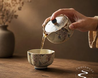 May include: A ceramic teapot with a scalloped rim and a matching teacup, both featuring a speckled cream glaze with hand-painted purple and green floral patterns. The teapot has a dark blue knob on the lid and is pouring tea into the cup.