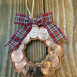 May include: A circular wreath ornament constructed from copper-colored United States one-cent coins arranged in a ring. The ornament is topped with a decorative red, green, and blue plaid fabric bow and includes a tan twine loop for hanging.